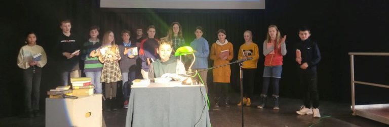 Schüler an Tisch auf Bühne vorlesend, mehrere Schüler dahinter stehend