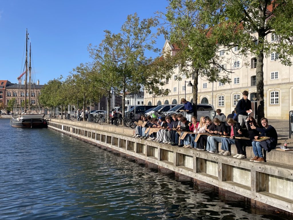 Gruppe Schüler mit Hr. Cornelius und Frau Meyborg Pizza essend am Wasser vor Stadtkulisse